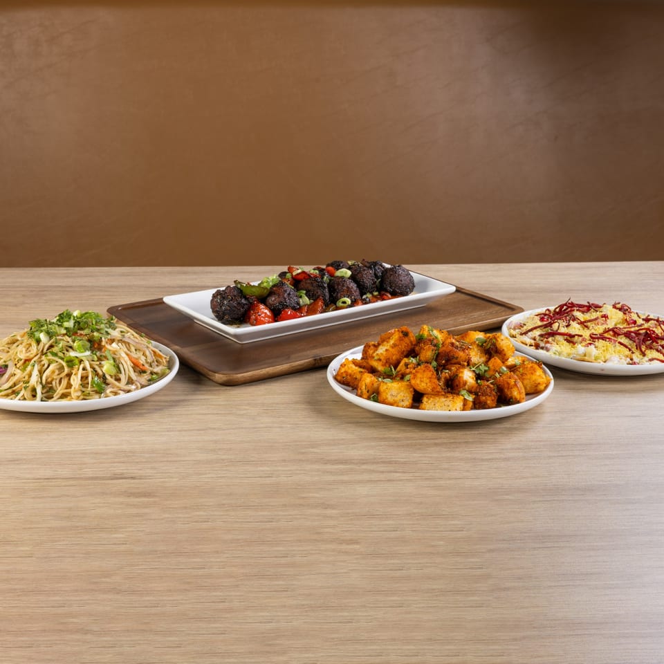 Four plates of Indian food, including noodles, fried paneer, and dark vegetable fritters, on a wooden table.