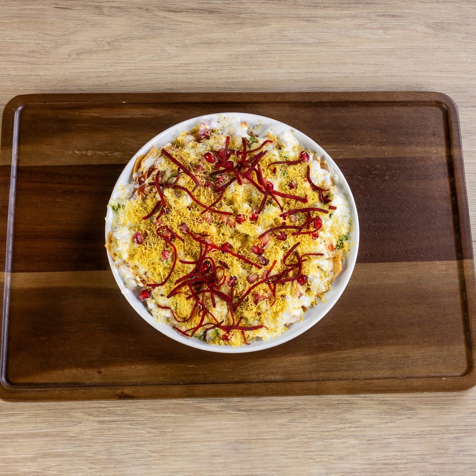 A white bowl of Indian Dahi Puri, topped with sev, pomegranate seeds, and beet juliennes.