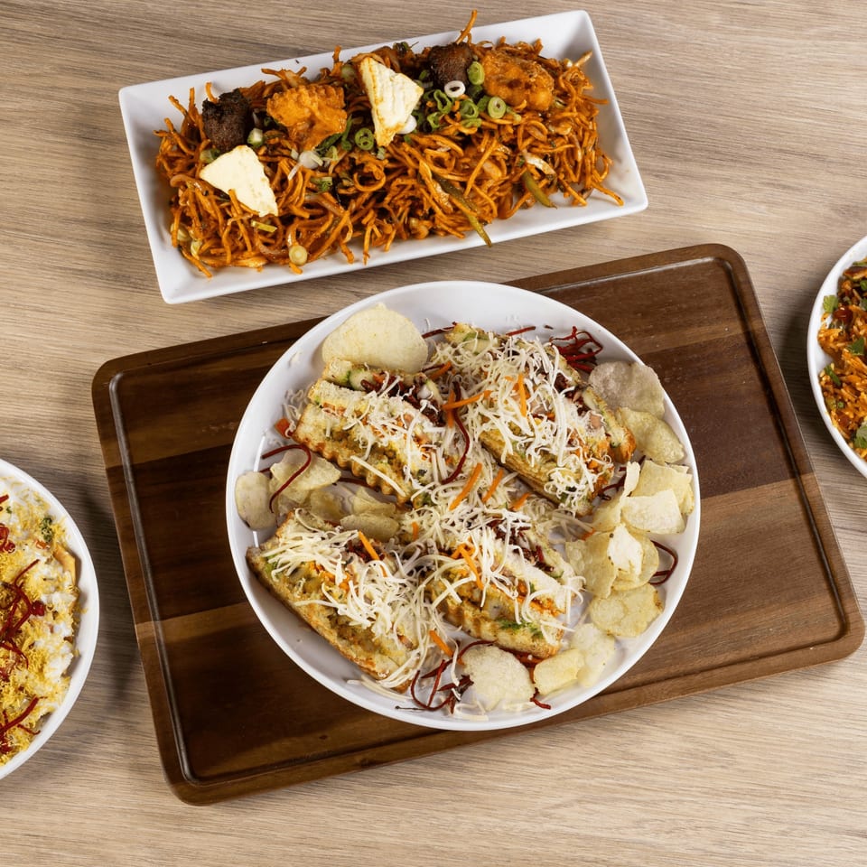 Overhead view of three Indian dishes: noodles with paneer, cheesy sandwiches with chips, and another dish.
