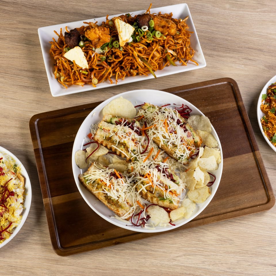 Overhead view of three Indian dishes: noodles, cheesy sandwiches with chips, and another dish with yellow toppings.