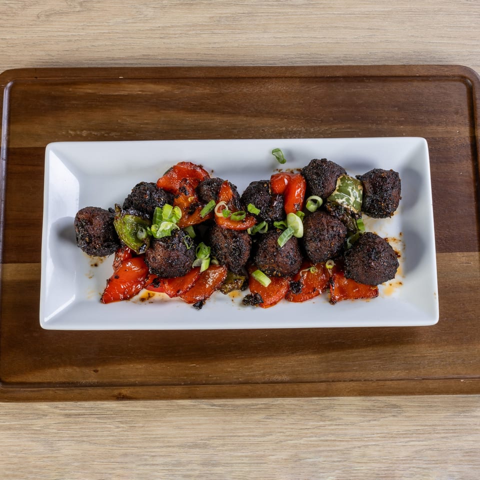 Indian dish with dark brown meatballs, red bell peppers, and green onions on a white rectangular plate.