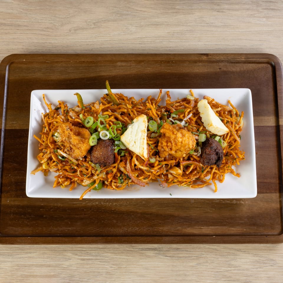 Indian noodles with fried paneer, vegetables, and dark brown pieces, served on a white rectangular plate.