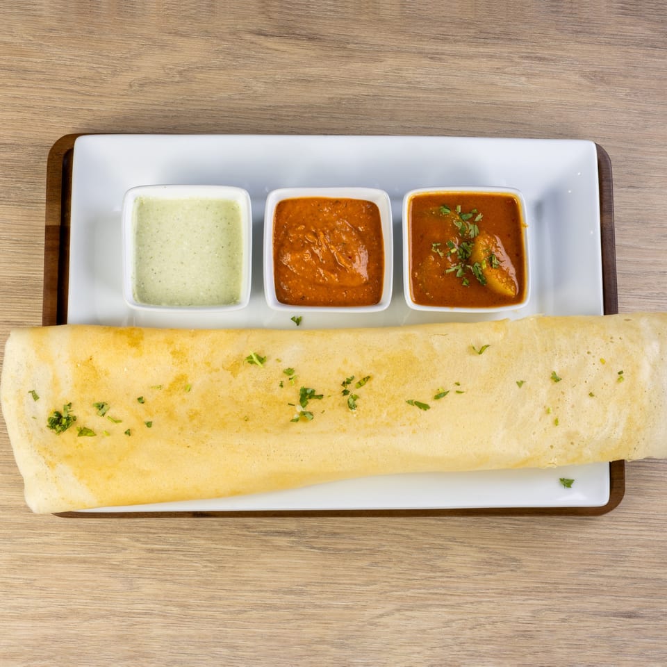 A long, golden-brown dosa with cilantro, served with three small bowls of Indian chutneys.
