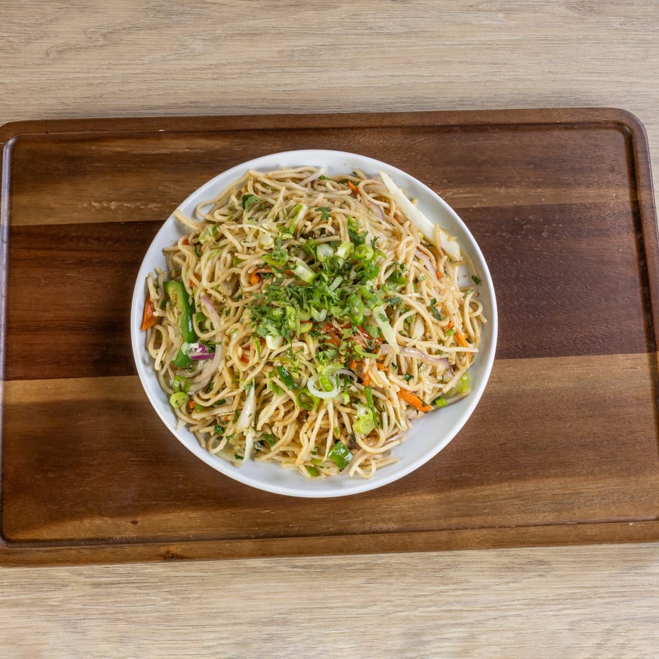 A white bowl of Indian-style noodles with vegetables and herbs on a wooden board.